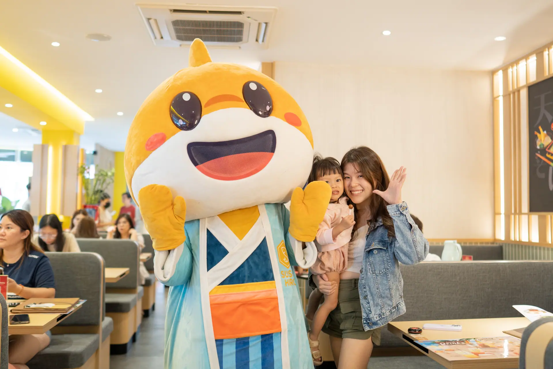 Hiro Boy waving with a mother and her toddler inside a Sushi Hiro outlet