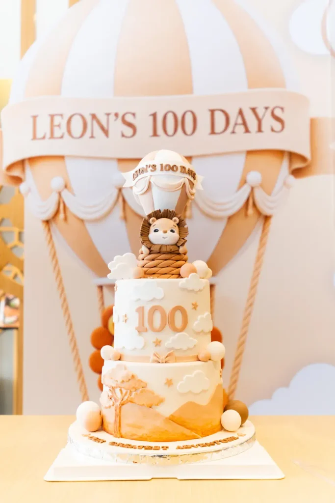 A tiered 100 days cake topped with a little lion in a hot-air balloon